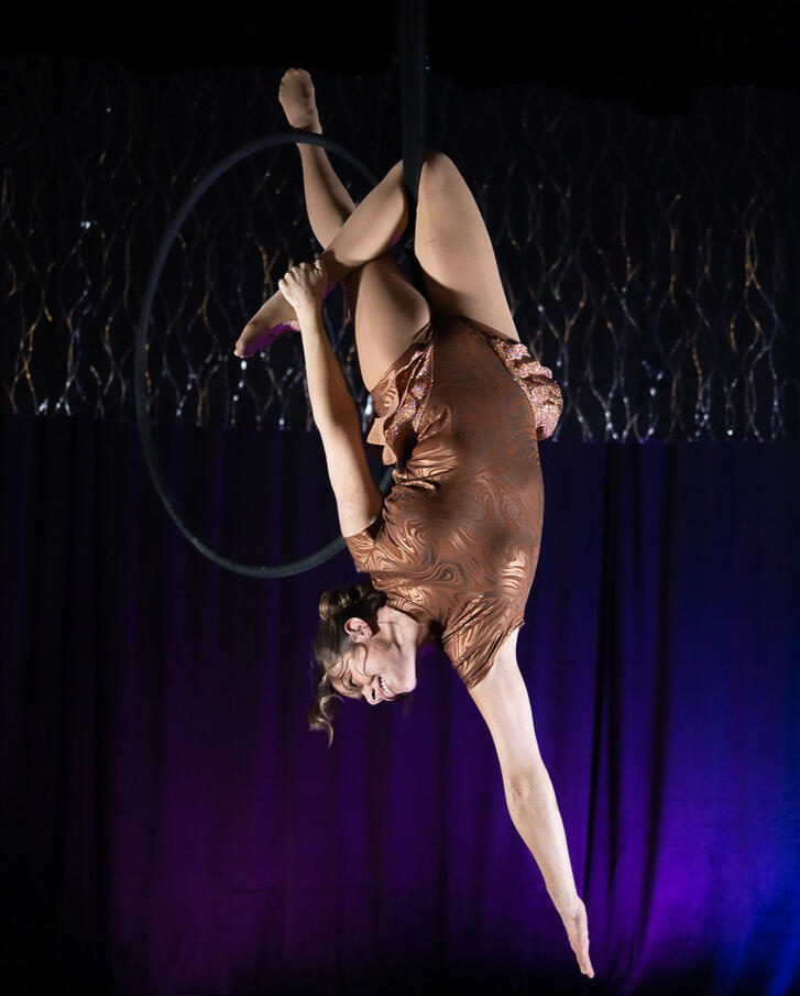 An aerialist wearing a tan leotard performs an advanced trick on a black aerial hoop