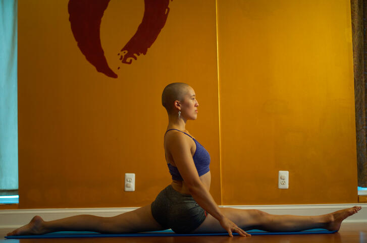 A bald woman wearing a purple sports bra and gray athletic shorts performs a front split in front of a gold wall